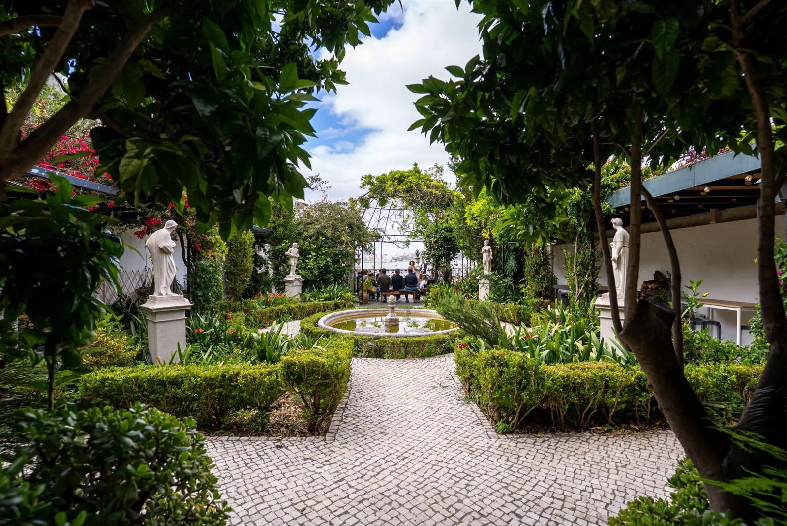 A cobbled path through a formal garden with statues and a fountain, leading to people gathered under a pergola with the river beyond