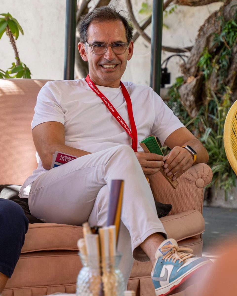 Gus smiling on a terrace sofa at Lisboa 2036, legs crossed, in a white T-shirt, white trousers and blue sneakers, with a red Fixed to Flow lanyard